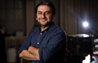 Tasan smailes with arms crossed in dark roo with spotlight shining in background on right.m