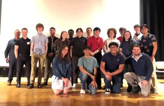 About 18 people, including teachers and students, stand on stage for a group photo.
