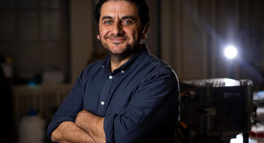 Tasan smailes with arms crossed in dark roo with spotlight shining in background on right.m